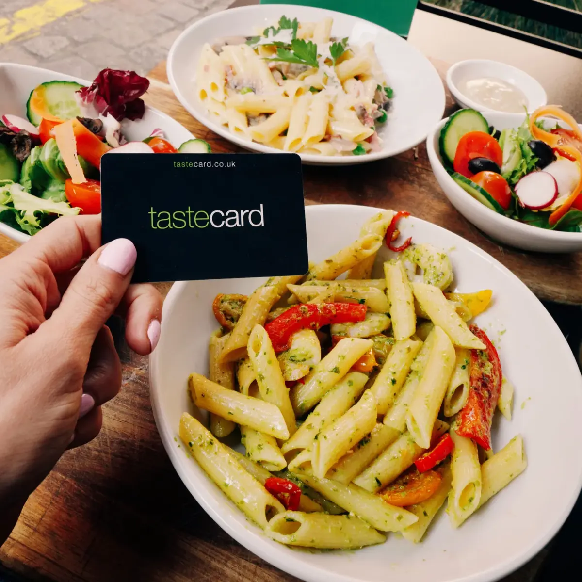 A hand holding a Tastecard above a bowl of pasta